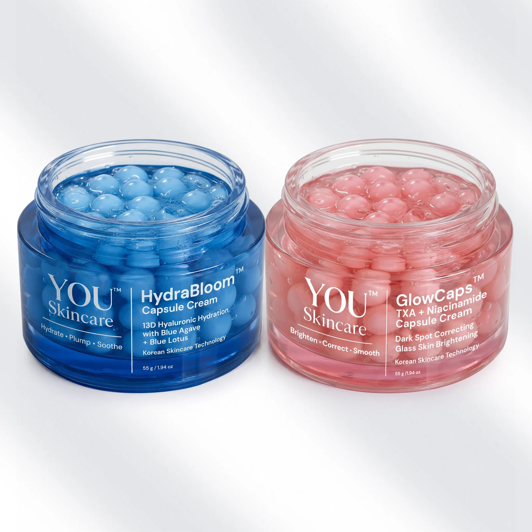 Two open cosmetic jars on a light gray background: a blue jar labeled YOU Skincare HydraBloom Capsule Cream and a pink jar labeled YOU Skincare GlowCaps TXA + Microneedle Capsule Cream, each filled with round gel capsules.