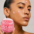 A close-up of a woman holding a jar of YOU Skincare GlowCapsule cream, with pink gel capsules visible inside the clear jar. The woman's face shows several small, raised pink gel capsules applied to her skin against a neutral light gray background.