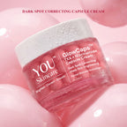 A jar of YOU Skincare GlowCaps TXA + Niacinamide Capsule Cream, shown in a pink translucent jar with a white lid, resting on a glossy pink background with pink spheres.
